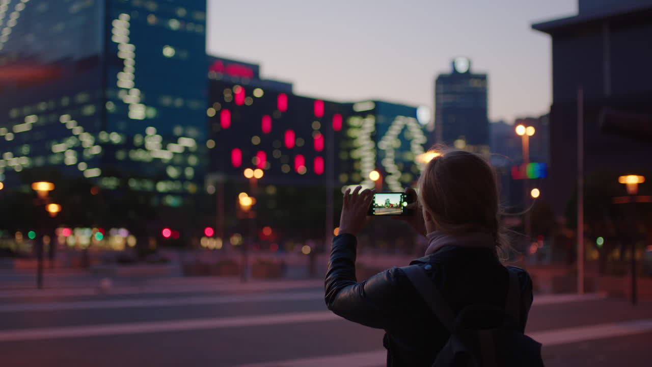 도시 저녁 관광을 즐기는 스마트폰 카메라를 사용하여 도시 조명 스카이라인의 사진을 찍는 젊은 금발 여성의 초상화