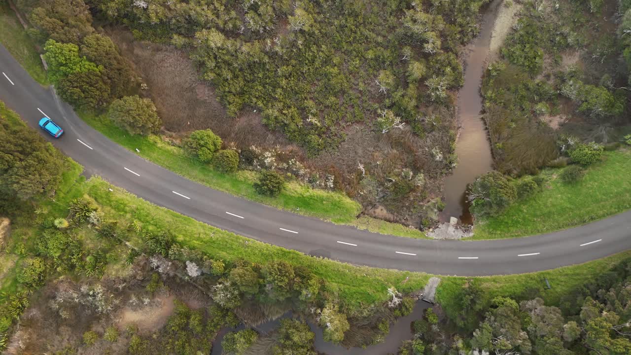 Blue car driving on winding country road through lush green landscape, scenic road trip adventure on Waiheke Island, New Zealand. Aerial top-down pov