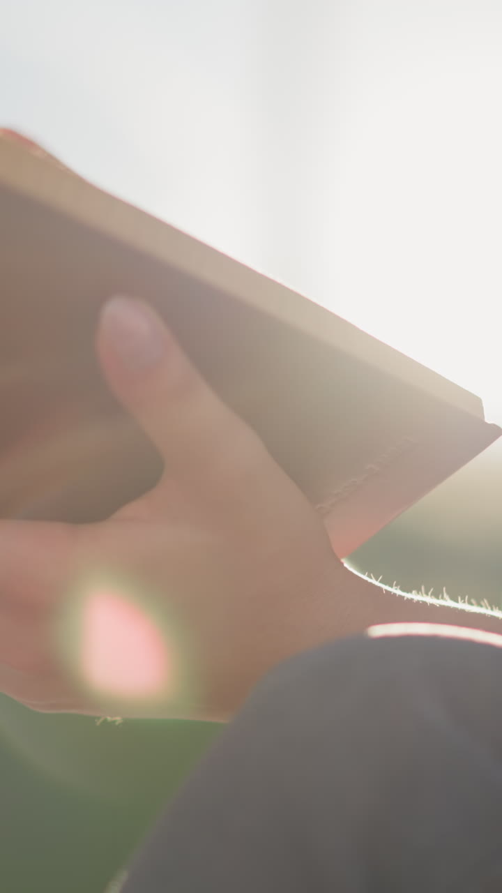 태양 광선은 밖에서 다리를 고 앉아있는 여성이 들고 있는 열린 책에 반사되며, 그녀의 모습은 배경에서 약간 흐릿해지고, 바람은 부드럽게 페이지를 통해 불고 있습니다.