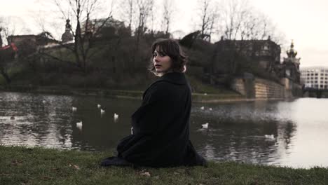 Junge-Frau-In-Einem-Schwarzen-Mantel,-Die-Im-Herbst-Am-Nachmittag-In-Einem-Park-Sitzt-Und-Enten-In-Einem-Meer-Füttert-3