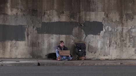 wide shot of a homeless veteran sitting on the sidewalk holding a cardboard sign asking for help