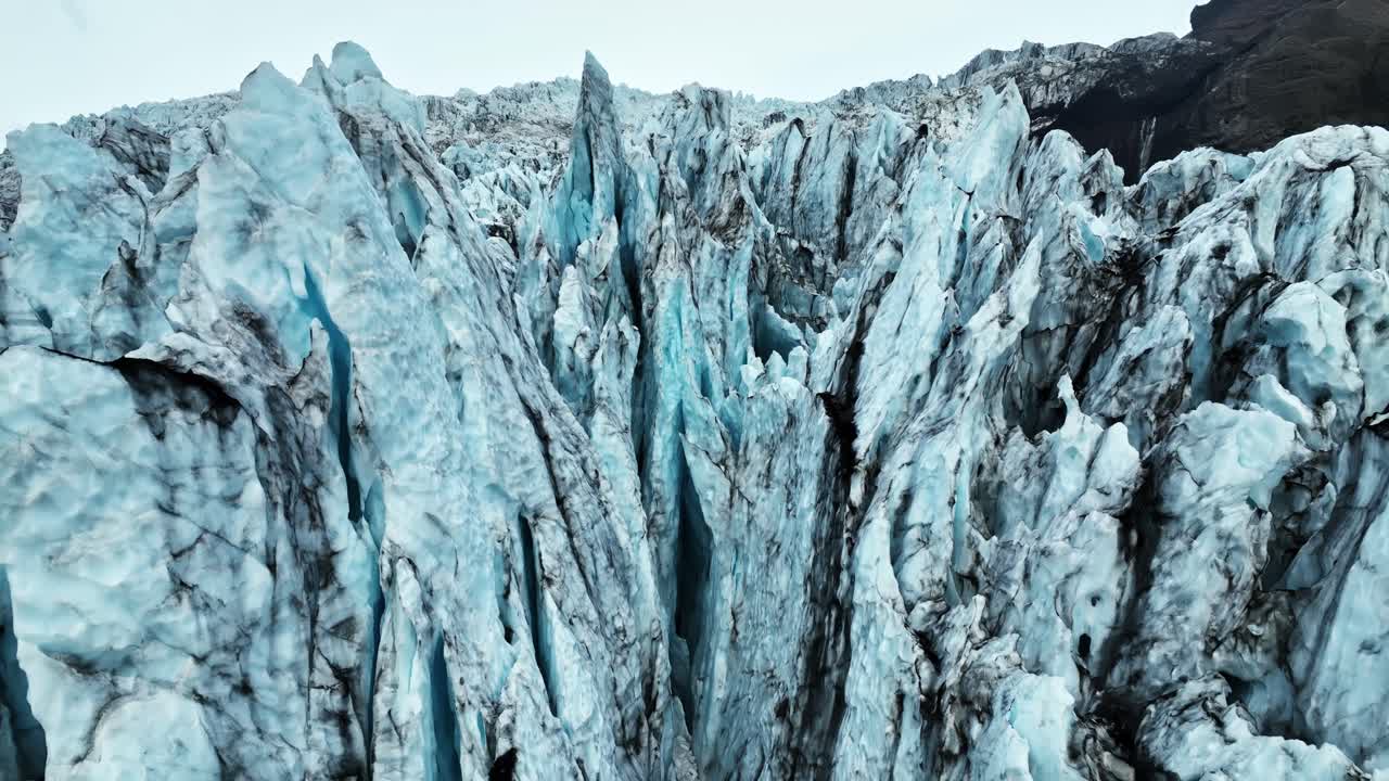 Un drone si avvicina a imponenti seracchi blu e profondi crepacci su un ghiacciaio islandese, segnato da striature scure di cenere vulcanica con un aspro sfondo montuoso