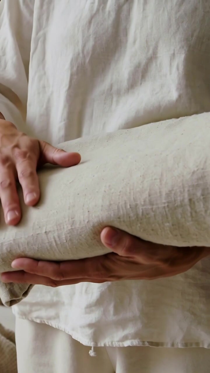Video verticale: Uomo che tiene delicatamente del lino, lisciando un rotolo in una stanza semplice, con una camicia larga, per appiattire le pieghe.