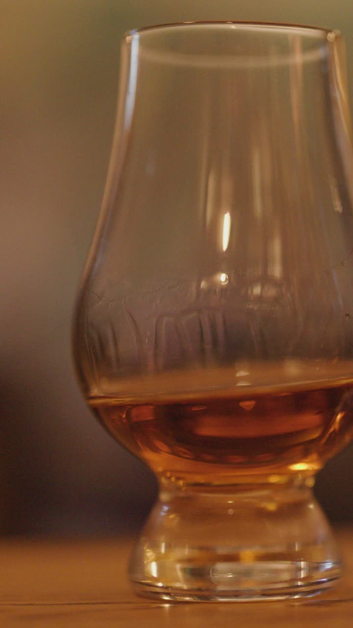 Una sequenza ravvicinata mostra whisky ambrato che vortica in un bicchiere da degustazione a tulipano, posizionato su un bancone di legno con illuminazione calda e soffusa e profondità di campo ridotta