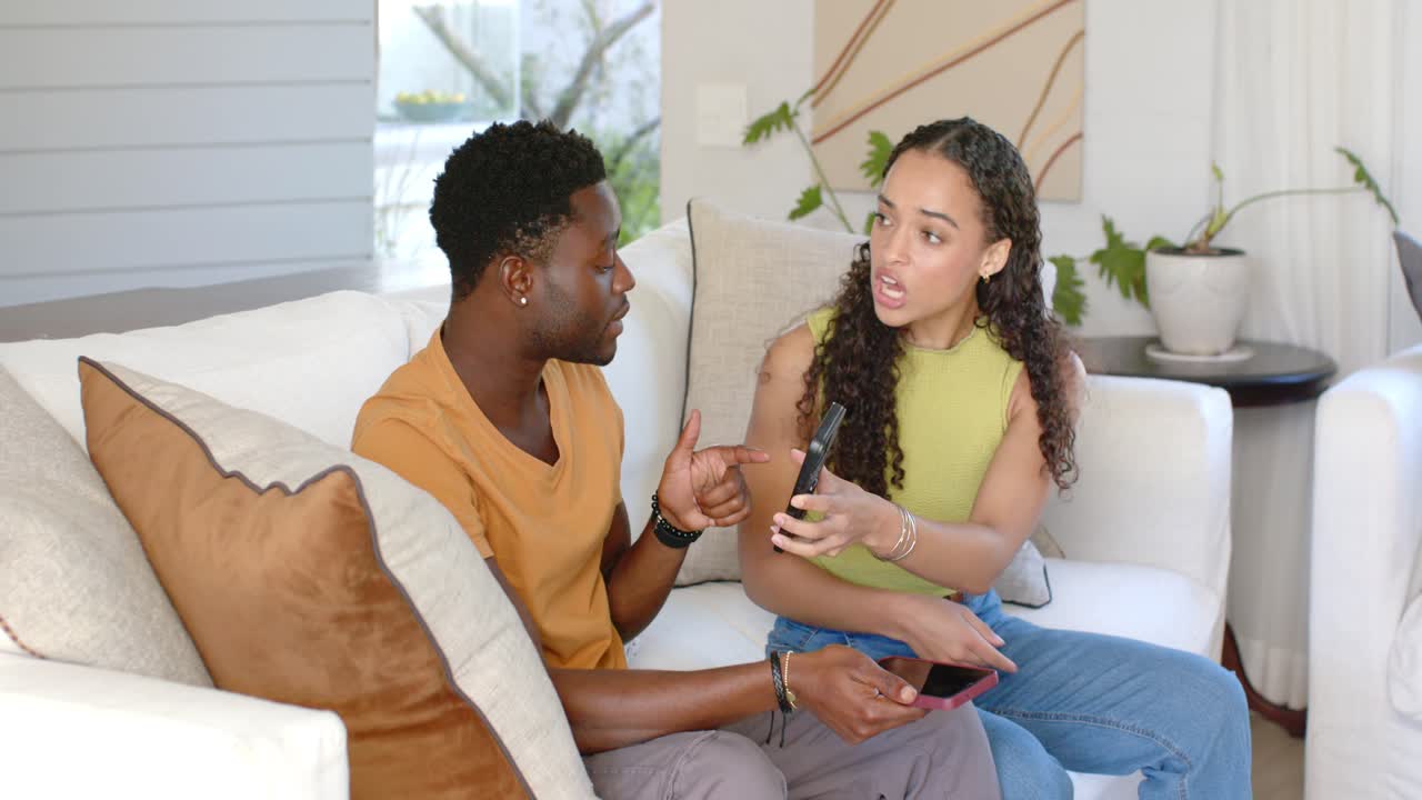 Couple sharing phones and remote, woman pointing phone and showing content on white sofa