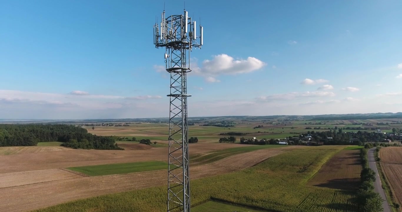 Aerial shot of an iron transmission tower surrounded by fields on a sunny summer day with blue skies and clouds, 4K