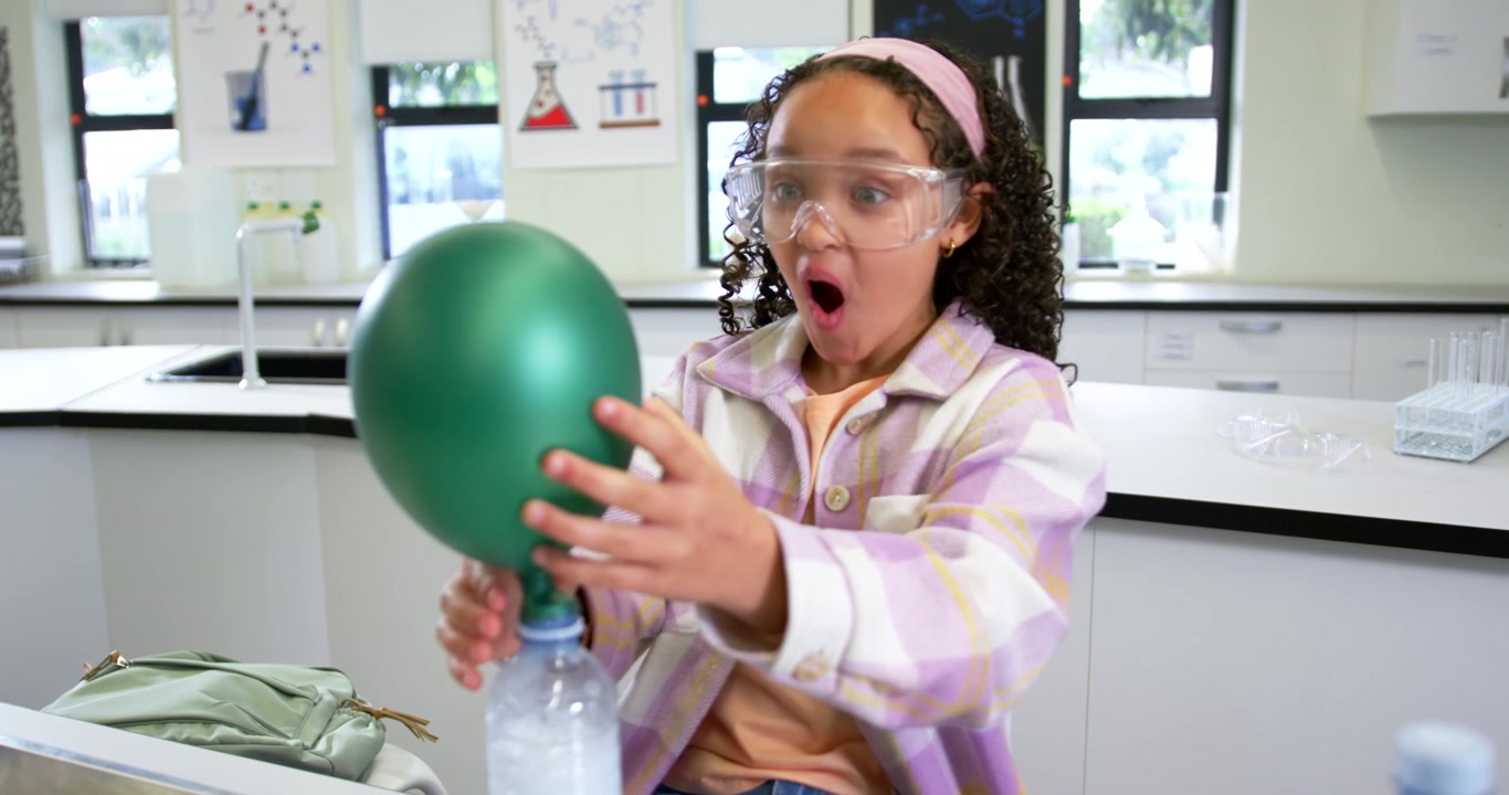 Preteen African American with goggles placing balloon on bottle at school lab, watching gas rising