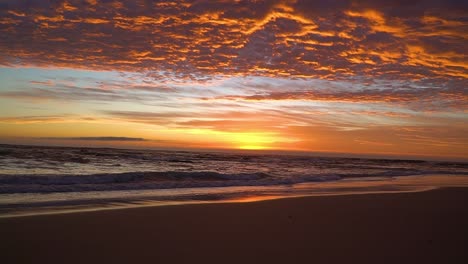 Zeitlupenfilm-Sonnenaufgang-Des-Feuerhimmels-Mit-Schäumenden-Meereswellen,-Die-Am-Goldenen-Reflexionsstrand-Des-Ufers-Plätschern