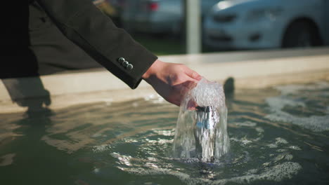 hand of a man in a black coat touching bubbling spring water, with blurred background featuring parked cars and outdoor greenery