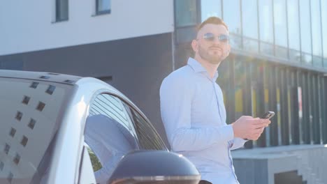 a rich man holds a smartphone in his hands while standing near his car he smiles and looks away a rich man holds a smartphone in his hands while standing near his car he smiles and looks away business man on the background of sun rays