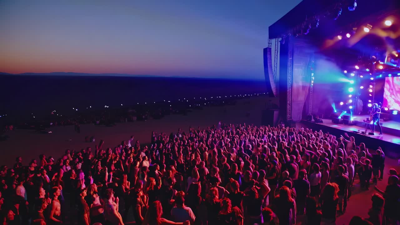 Large group of people attending a live music concert at an outdoor venue on the beach, enjoying the music and atmosphere during a vibrant sunset