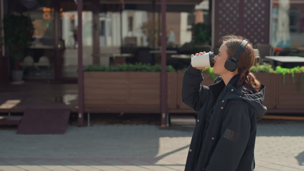 Donna caucasica che cammina per le vie della città sorseggiando un caffè mentre indossa cuffie e un cappotto nero, sorriso rilassato, passa davanti al dehors di un bar con fioriere, luce solare mattutina soffusa e bokeh delicato, spontanea
