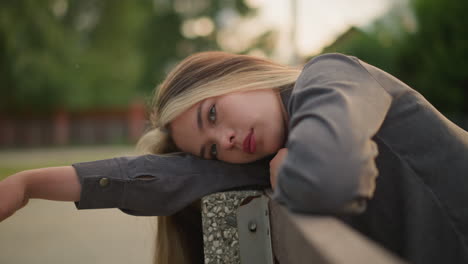 close-up of woman in grey clothing resting head on arm outdoors, lost in thought with a calm expression, background is blurred featuring greenery