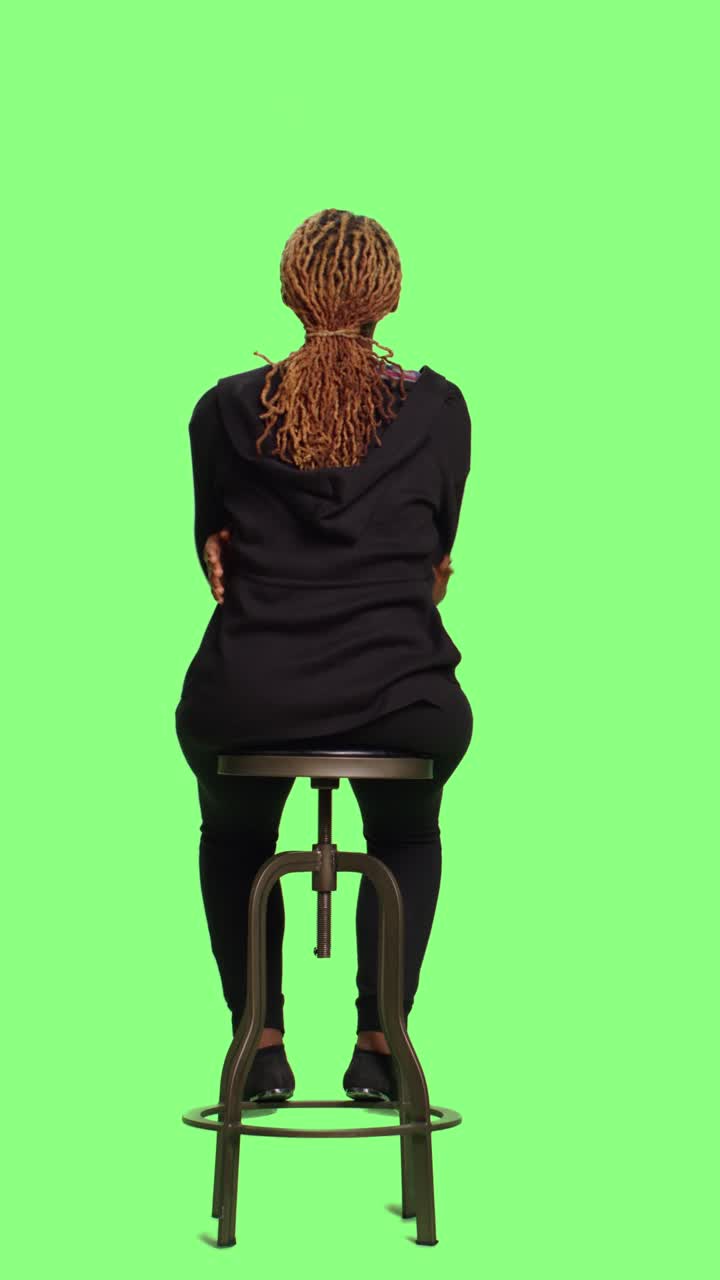 Video verticale: ragazza afroamericana seduta su una sedia su greenscreen a figura intera