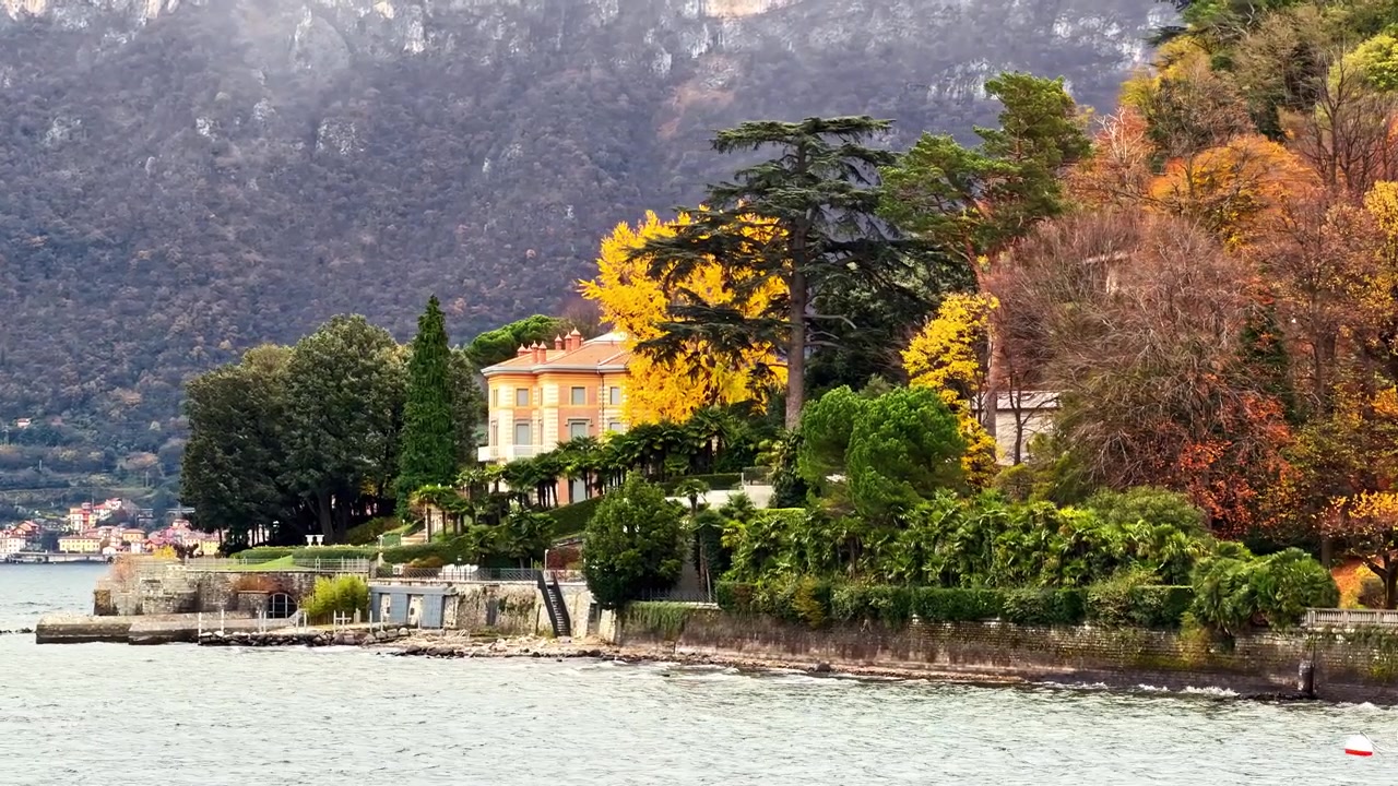 Drone aereo che si sposta a sinistra sopra un'elegante villa sul lago e giardini autunnali colorati sulle rive del Lago di Como a Bellagio, incarnando la bellezza senza tempo dell'autunno lacustre italiano