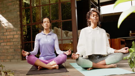 two multiracial female friends meditating on yoga mats on porch, tranquility and mindfulness