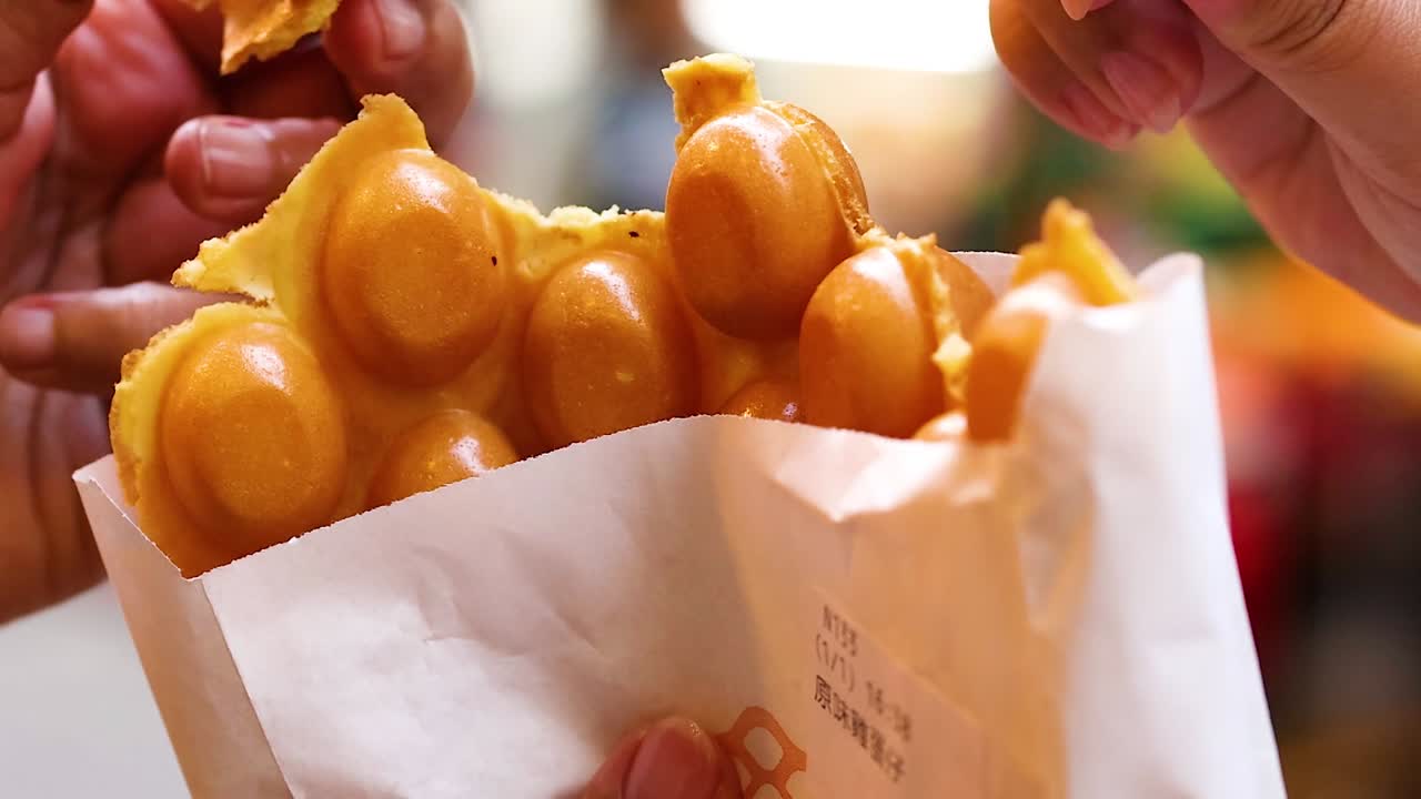 Close-up of hands tearing bubble waffles from a paper bag, showcasing texture and enjoyment.