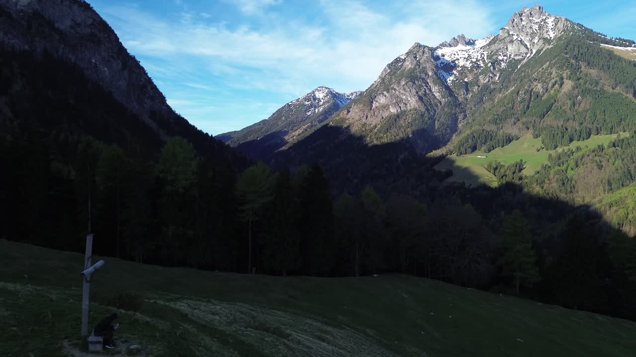 Eine Person mit schwarzer Jacke sitzt im Schatten neben einem Gipfelkreuz und fliegt an einem sonnigen Tag in Österreich, Europa, eine Drohne in der Berglandschaft.