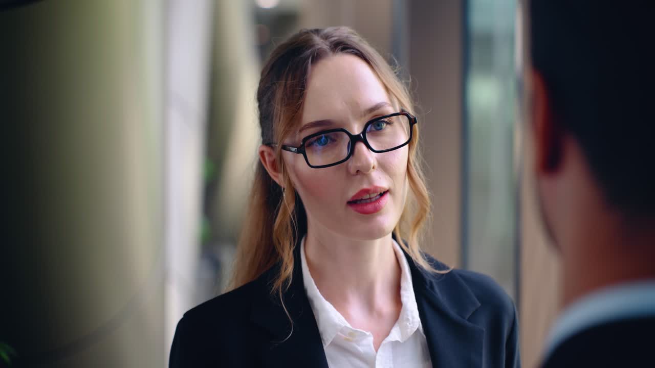 Face close up of serious businesswoman with glasses talking intently to male colleague about work problems while standing near windows in modern corporate office