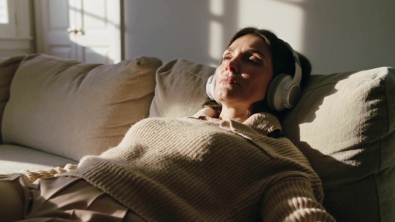 Reclining woman in knit sweater on sofa adjusting headphones near window, closing eyes listening