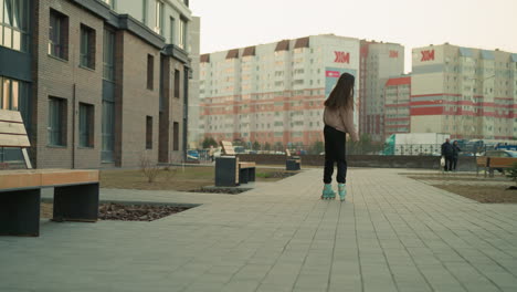 una foto trasera de una joven patinando a lo largo de un camino pavimentado en un parque urbano. ella lleva una chaqueta de melocotón y pantalones negros, altos edificios residenciales visibles en el fondo