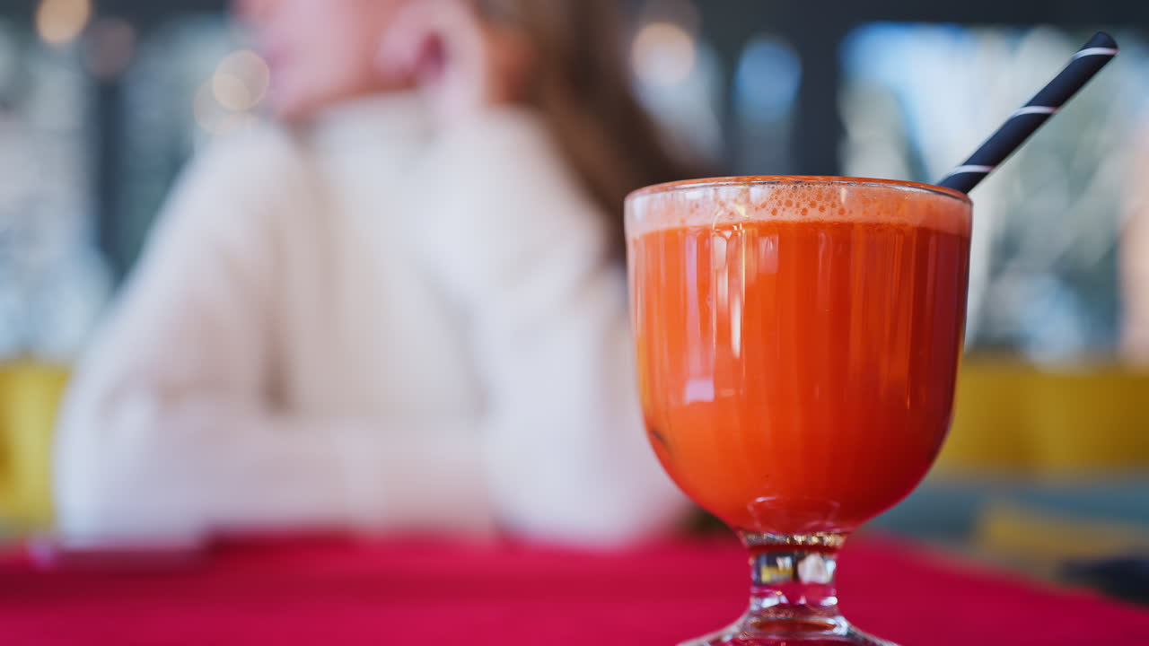 Close up of an orange and carrot juice in a glass with a black straw on a red table cloth with a woman on the background