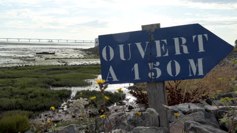 Blue-wooden-sign-with-a-distance,-in-the-rear-of-the-bridge-to-leave-the-island-of-Oleron,-France