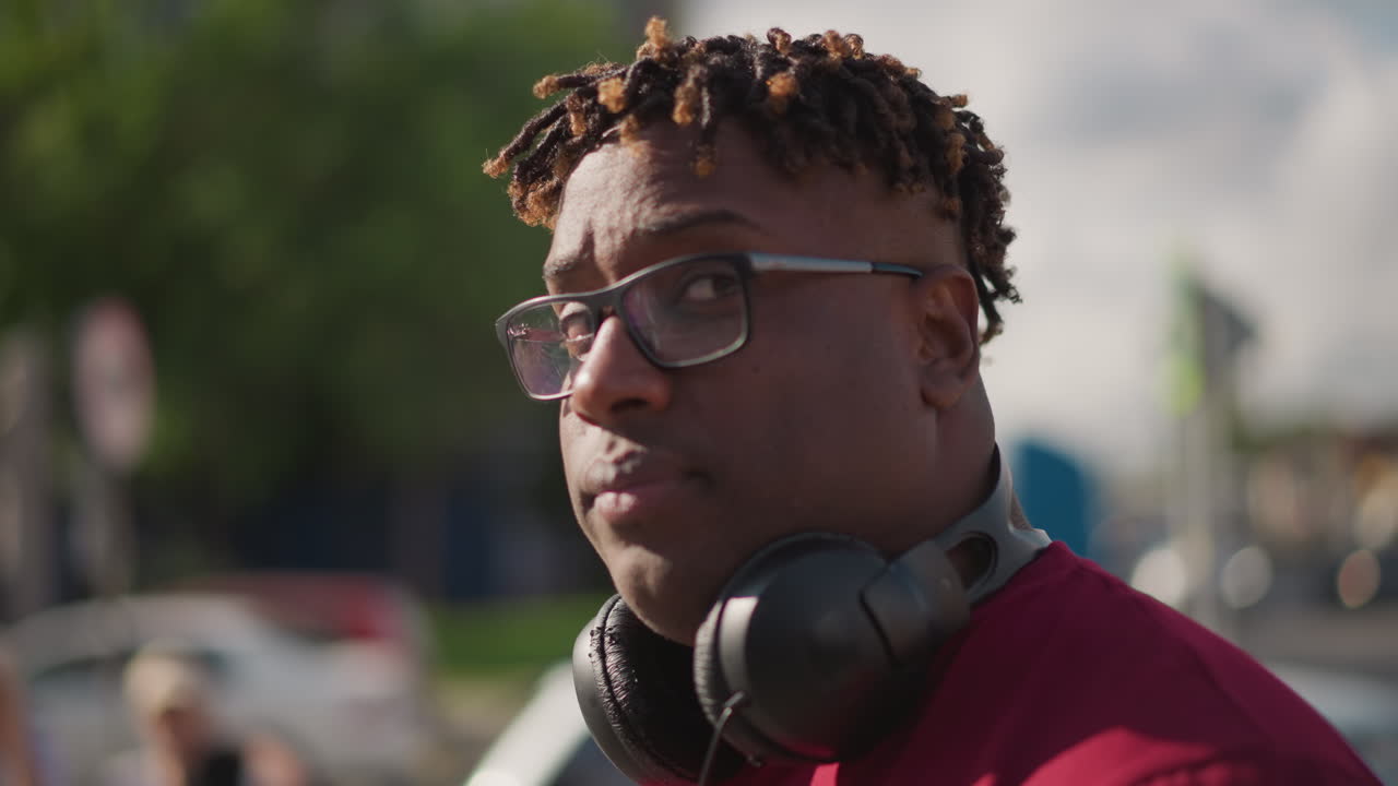 DJ nero con cuffie per strada, musicista all'aperto che ascolta e sorride indossando occhiali e dreadlocks, sfondo diurno della città con profondità di campo ridotta, ritratti ravvicinati spontanei che catturano l'attimo
