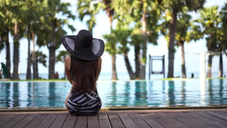 Eine-Junge,-Stilvolle-Frau-In-Einem-Schwarz-weißen-Badeanzug-Sitzt-Auf-Der-Holzterrasse,-Die-Einen-Resort-Pool-Umgibt-Und-Auf-Das-Meer-Blickt