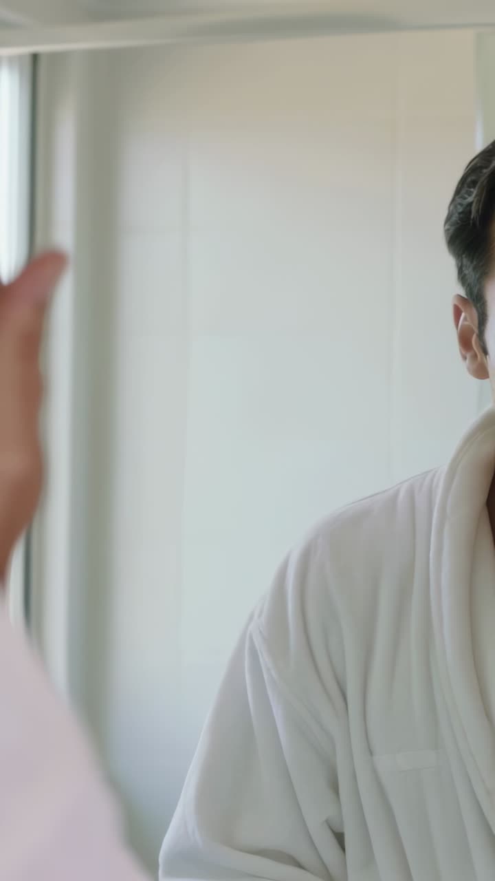 Vertical video: Noticing mirror, man in robe adjusting under-eye patches in bathroom, checking fit