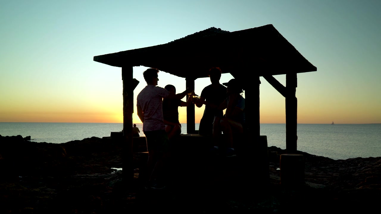 Silhuetter av venner som skåler med ølglass ved strandgrillen i solnedgangen.
