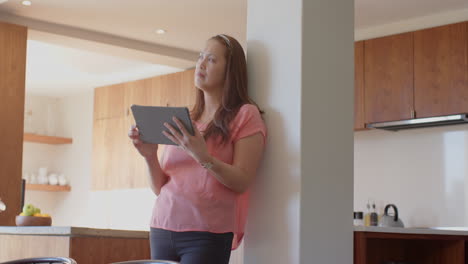 using tablet, woman standing in kitchen, looking thoughtfully out window