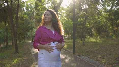 pregnant woman expectant mother walking alone in park, attractive caucasian casual dressed mom stroking caressing touching her belly with hands relaxing, childbearing tenderness, medium portrait shot