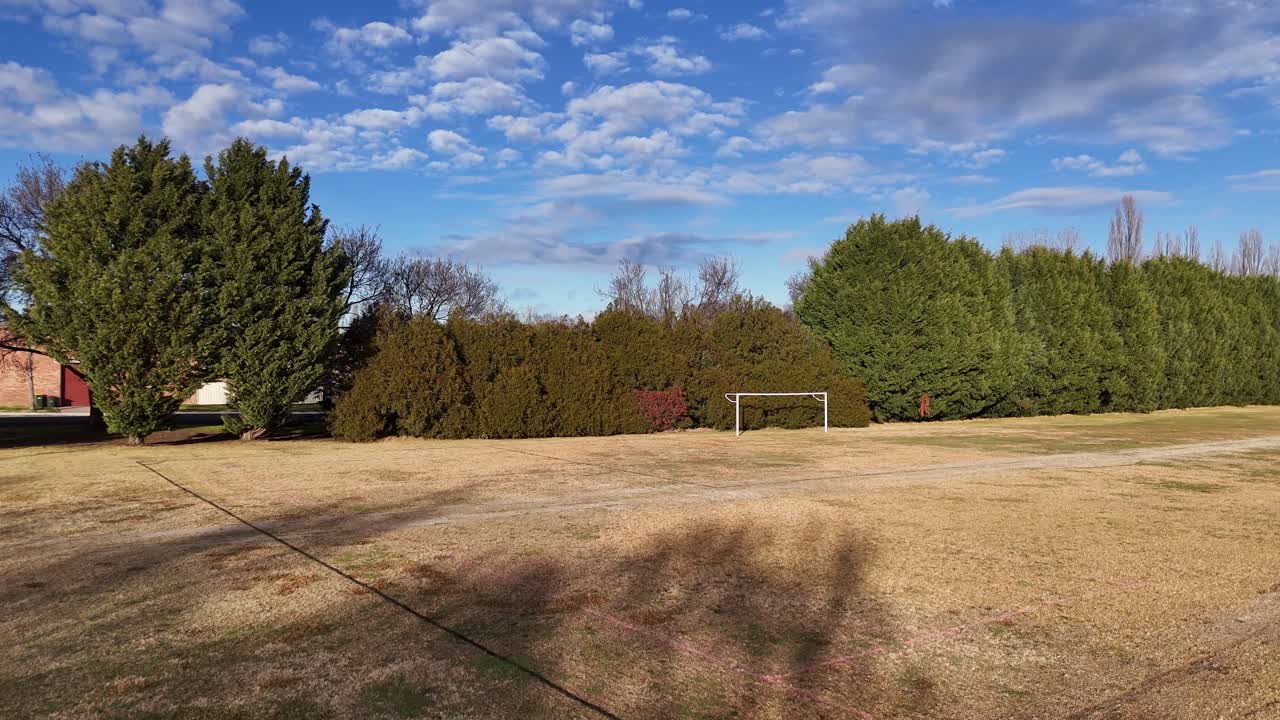 Static camera captures an empty sports ground with a soccer goal, dry grass, and scattered trees in Glen Innes, Australia, under bright daylight