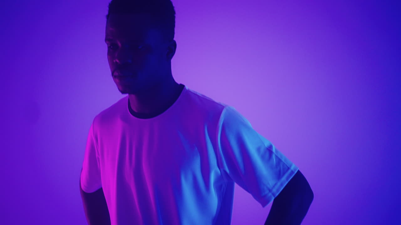 retrato en cámara lenta de un jugador de fútbol profesional negro en un uniforme blanco mirando a la cámara en la luz de neón azul-rojo del estudio. un jugador de fútbol brutal confiado y victorioso