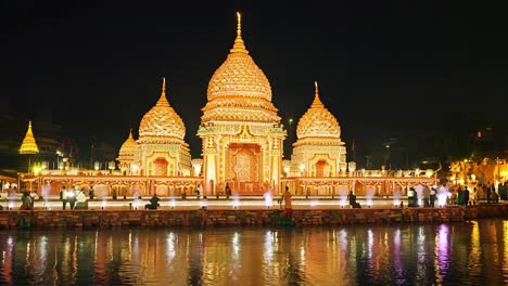 tourists gather by the water, admiring a beautifully illuminated traditional thai temple at night. colorful lights enhance the stunning architecture, creating a vibrant atmosphere