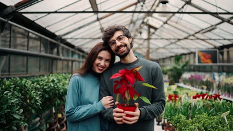 Beautiful-young-couple-in-casual-clothes-is-holding-a-plant,-looking-at-camera-and-smiling