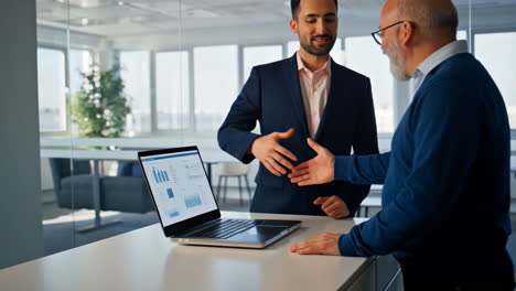 businessmen shaking hands in a modern office with laptop displaying charts