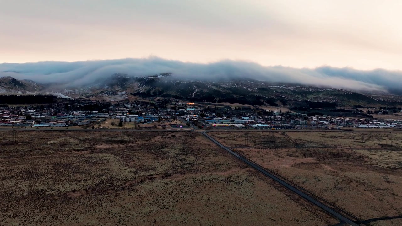 Drone-hyperlapse over Hveragerdi på Island som viser lave skyer som beveger seg over fjellene, med utsikt over byen, landskapet og det omkringliggende terrenget.