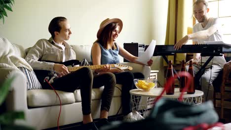 young people popular band are holding rehearsal at home, singing and playing the keyboard and the guitar and checking paper with lyrics and music sheet.