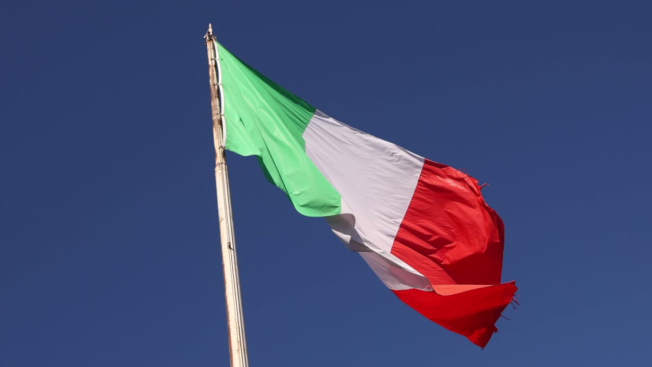 Primo piano della bandiera nazionale italiana che sventola orgogliosamente al vento contro un cielo azzurro limpido.
