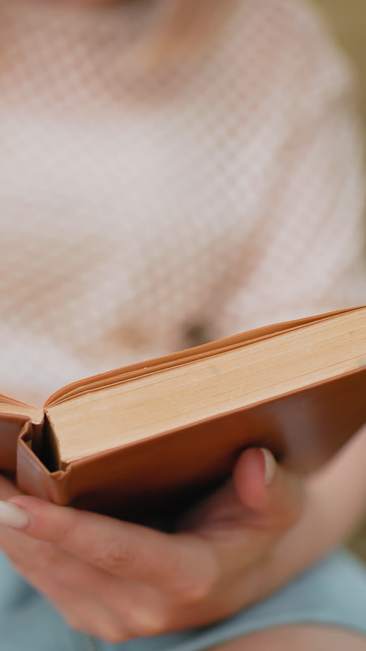 야외에서 책을 읽는 동안 열린 책에서 페이지를 뒤집는 여성의 클로즈업, 파란색 쇼트와 섬세한 손에 부드러운 초점, 자연스러운 흐릿한 배경과 함께 연구와 반성의 평화로운 순간