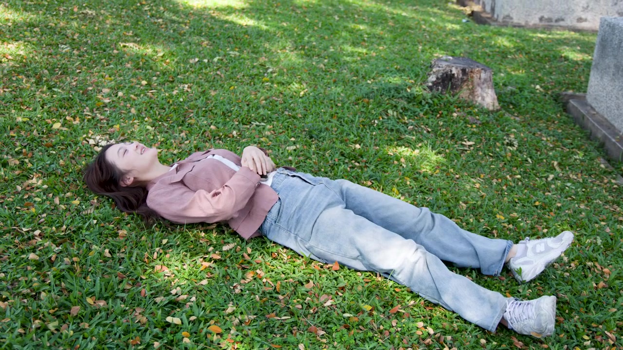 A young Asian woman sits on campus grass, adjusts her hair, and slowly lies back