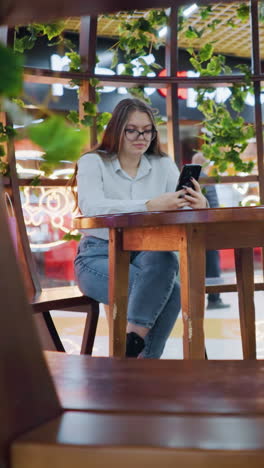 mujer joven chic sentada en un sentado de madera decorativa, sosteniendo el teléfono, bolsas de compras a su lado, transeúnte caminando en un vibrante y bullicioso centro comercial con luces de fondo dinámicas