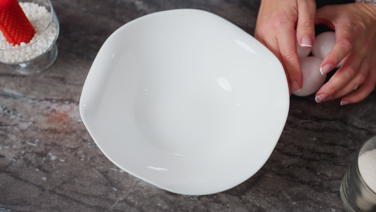 Aerial view of chef hand placing white bowl on kitchen table, gently dropping eggs while wearing a bracelet, red burning candle and glass cup of sugar on countertop