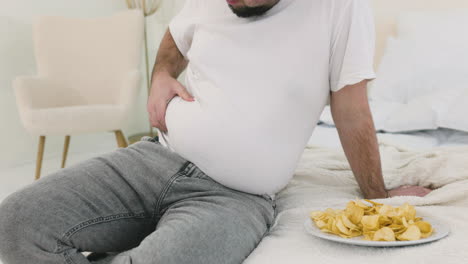 niño tocándose la grasa del vientre sentado en la cama cerca de papas fritas