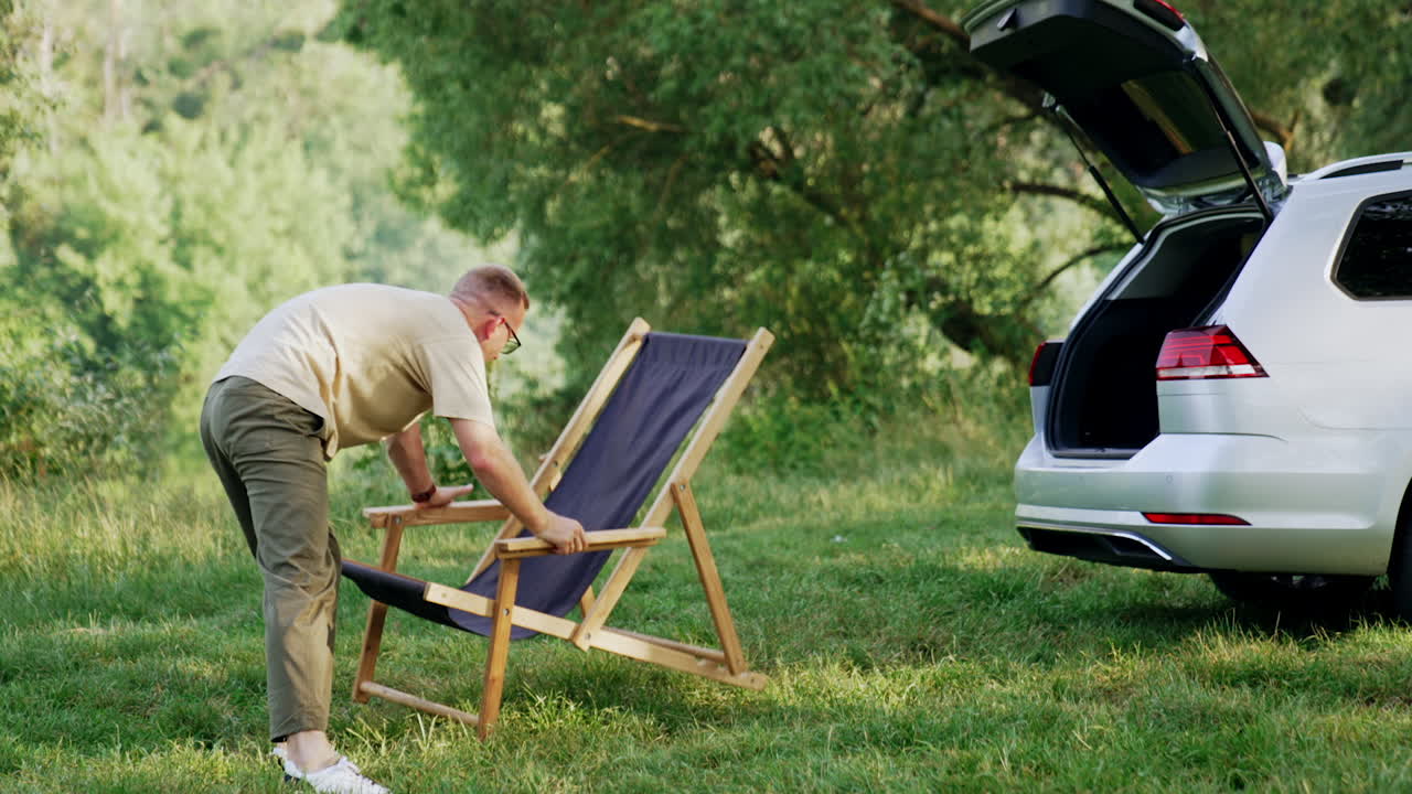 Caucasian man sets folding chair on the grass. Man chooses the best place for the chair. Nature at backdrop.