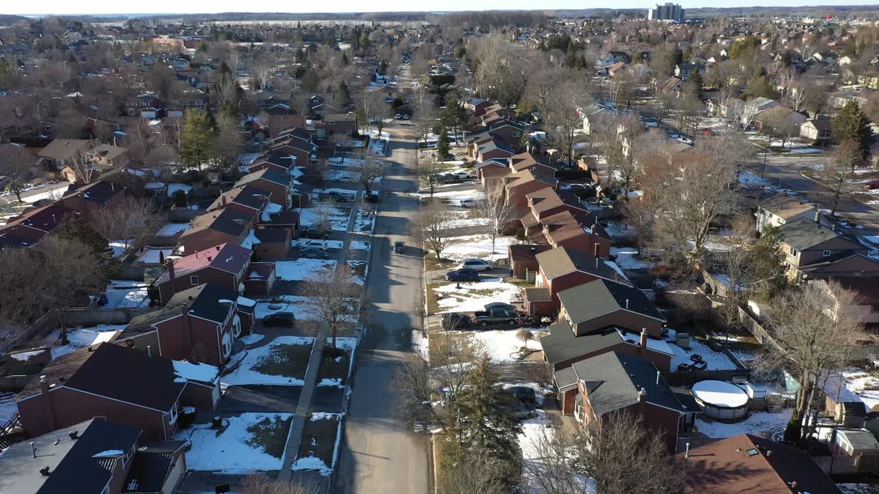 Aerial view flying slowly over a residential street in a suburban neighborhood during winter, with snow on the ground and rooftops, and barren trees lining the quiet road in the morning sun