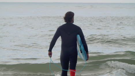 back view of a man with artificial leg in wetsuit and surfboard walking into the ocean