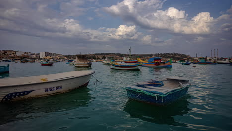 Local-Maltese-boats-docked-near-island-shore,-time-lapse-view-with-moving-clouds